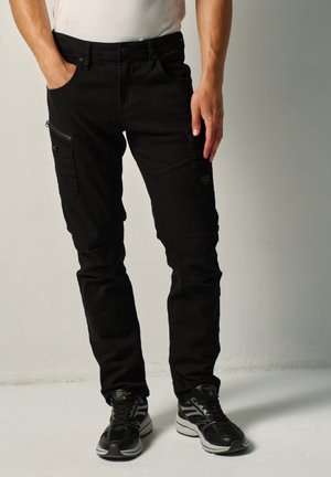 Hombre con pantalones cargo negros con bolsillos con cremallera y solapa, combinado con zapatillas negras y blancas, de pie con una mano en el bolsillo.
