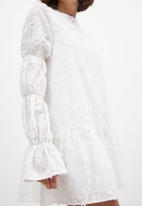 Balta siuvinėjimo suknelė su gėlių raštu, ilgos pūstos rankovės su elastinėmis apykaklėmis ir plačiomis volanomis apačioje.