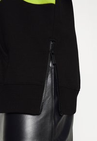 Sudadera negra con un acento verde, presenta un zipper lateral y un dobladillo acanalado. El material es suave y se complementa con una falda de cuero brillante por debajo.