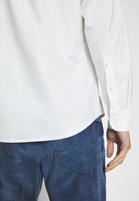 Person i långärmad vit skjorta och blå kordflamsbyxor, sedd från ryggens midja till handen på en plain bakgrund.