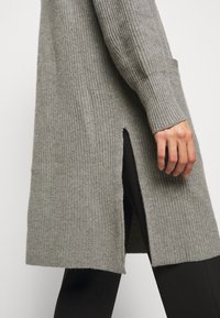 Grå stickad lång cardigan med sidoförslitning, bärs över svarta byxor, visar ärmar i ribbat mönster som når till armbågen samt en delvis framficka.