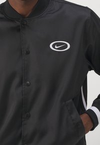 Černá bunda vyrobená z hladké tkaniny, která má patentky a bílé logo Nike. Na rukávech jsou kontrastní bílé pruhy.