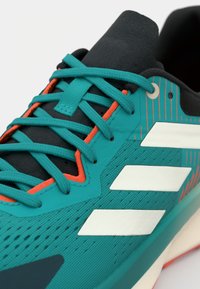 Türkisfarbiger Sportschuh mit weißen Streifen, schwarzer Zunge und türkisfarbenen Schnürsenkeln, die durch orange und türkisfarbene Ösen geführt sind.