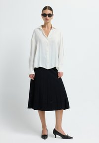 Chemise blanche à boutons avec une coupe décontractée, associée à une jupe midi plissée noire. Des escarpins noirs à bout pointu complètent la tenue.