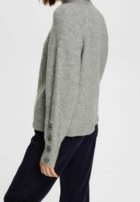 Pull en tricot gris avec un col montant et une texture côtelée. Éléments de boutons sur la manche gauche et coupe décontractée.