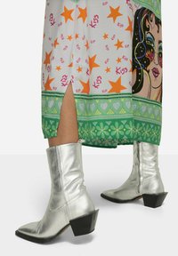 Silberne metallische Stiefeletten mit spitzer Zehe und chunky schwarzer Absatz, kombiniert mit einem bunten gemusterten Rock, der orangefarbene Sterne und grüne Akzente aufweist.