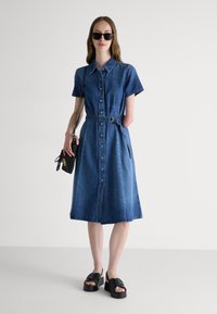 Denim shirt dress with short sleeves, front buttons, belted waist, and a flared skirt. Paired with black platform sandals and a small bag.