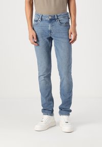 Jasnoniebieskie jeansy o slim fit z pięcioma kieszeniami i kontrastowymi przeszyciami. Noszone z białymi sneakersami i dopasowanym beżowym t-shirtem.