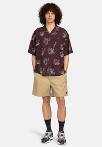 Burgunderfarbendes Kurzarmhemd mit einem Tiermotiv, kombiniert mit beigen Shorts. Das Model trägt dunkle Socken und mehrfarbige Sneaker.
