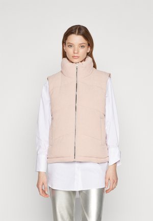 Chaleco acolchado en suave tela rosa claro con cuello alto y cremallera frontal. Presenta costuras horizontales y diseño sin mangas. Llevado sobre una camisa blanca.
