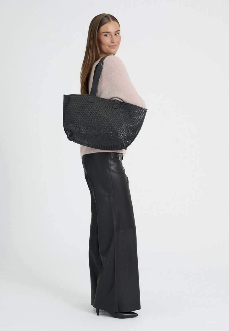 Femme portant un pull beige et un pantalon large noir tient un grand sac à main noir tissé sur l'épaule, souriant par-dessus son épaule.