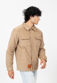 Kurtka z sztruksu w kolorze khaki z kołnierzem, zatrzaskami, dwoma przednimi kieszeniami oraz boczną kieszenią na zamek. Posiada pomarańczową metkę przy dolnym brzegu.