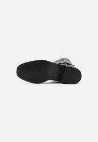 Bota de cuero negra con una suela texturizada, detalle de hebilla lateral y acabado suave. Presenta una puntera redondeada y un tacón bajo.