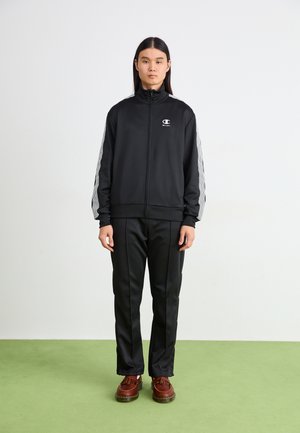 Chaqueta deportiva negra con cremalleras y rayas grises en las mangas, combinada con pantalones negros. El modelo lleva zapatos marrones con detalles texturizados.