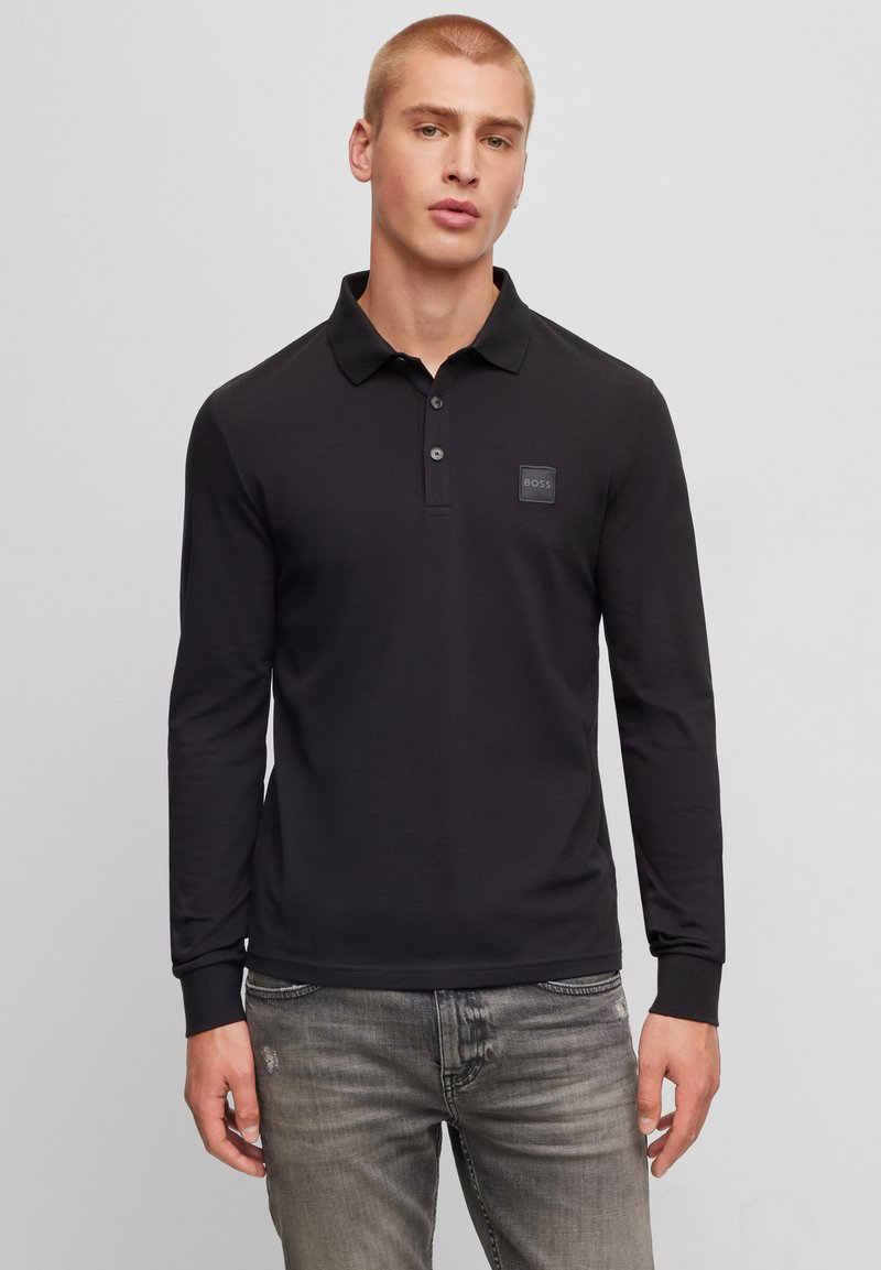 Långärmad svart polo med en placket med tre knappar och en texturerad logotyp på bröstet, ihop med slitna grå jeans.