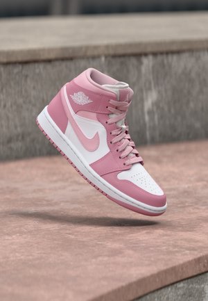 Ore viduryje pakibęs rožinės ir baltos spalvos aukštos Air Jordan sportinio batelio modelis su raišteliais, eksponuojamas prieš betono foną.