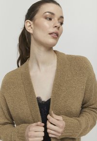 Brun stickad cardigan med strukturerad yta, öppen fram, och långa ärmar, bärs över en svart spets top.