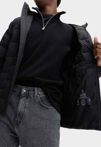 Giacca imbottita nera con collo zip, indossata sopra una maglietta nera e jeans grigi. L'interno presenta istruzioni per la cura e dettagli sui materiali.