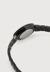 Μαύρο μεταλλικό ρολόι χειρός με συνδεδεμένο μπρασελέ, πλευρική όψη που δείχνει το κουρδιστήρι με το λογότυπο "ck" και πίσω κάλυμμα από ανοξείδωτο ατσάλι σε λευκό φόντο.