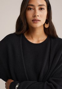 Juodas per didelis megztinis su apvalia iškirpte ir tekstūruotu audiniu. Turi ilgus rankovėmis ir laisvą siluetą, pabrėžtas dideliais kabučiais auskarais.