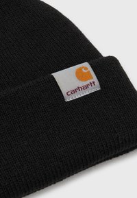 Zwarte gebreide beanie van zachte wol, met een omgeslagen rand. Een rechthoekig label toont een oranje logo en bordeauxrode tekst.