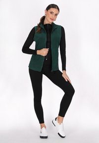 Grüne Fleece-Weste mit hohem Kragen, Reißverschluss und seitlichen Taschen, kombiniert mit einem schwarzen Langarmtop und schwarzen Leggings, abgerundet mit weißen Schuhen.