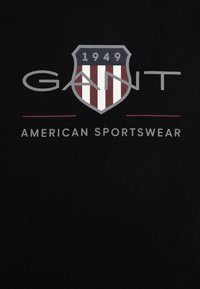 Svart bomullströja med en stor logotyp i skölddesign, röda och vita ränder samt texten "GANT AMERICAN SPORTSWEAR."