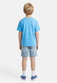 Παιδί με κοντά ξανθά μαλλιά που φορά φωτεινό μπλε μπλουζάκι, ανοιχτόχρωμο τζιν σορτς, λευκές κάλτσες και μπλε παπούτσια χωρίς κορδόνια, στέκεται γυρισμένο με την πλάτη.