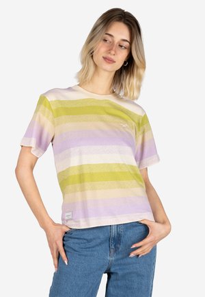 Frau trägt ein pastellgestreiftes T-Shirt mit gelben und lavendelfarbenen Streifen, posiert mit den Händen auf den Hüften in blauen Jeans vor einem einfarbigen Hintergrund.