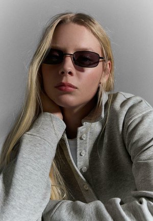 Giovane donna con capelli biondi lunghi che indossa occhiali da sole rettangolari scuri e un maglione grigio chiaro con bottoni, appoggiando la testa sulla mano.