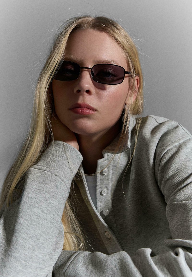 Jeune femme aux longs cheveux blonds portant des lunettes de soleil rectangulaires foncées et un pull boutonné gris clair, reposant sa tête sur sa main.