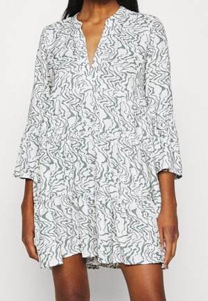 Kobieta ubrana w białą sukienkę z długim rękawem, wielowarstwową, z szarym, falistym, abstrakcyjnym wzorem i dekoltem w kształcie litery V.