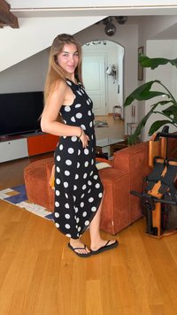 Νέα γυναίκα που χαμογελάει, φοράει μαύρο φόρεμα με λευκές πουά και σαγιονάρες, στέκεται σε ένα σαλόνι με πορτοκαλί έπιπλα και ένα φυτό.