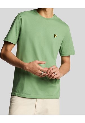 Uomo che indossa una t-shirt verde pastello a maniche corte con piccolo logo giallo di uccello sul petto e pantaloni color avorio, mani insieme che mostrano anelli d'argento.