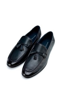 Mocasines de cuero negro con textura suave, que presentan un borlas decorativa y costuras en contraste. Punta redondeada y diseño de perfil bajo.