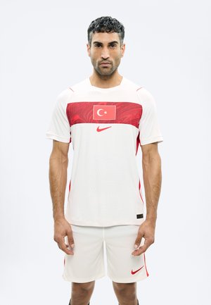Homme debout, face à l'avant, portant un uniforme de sport blanc et rouge avec le drapeau turc et le logo Nike sur la poitrine et le short.