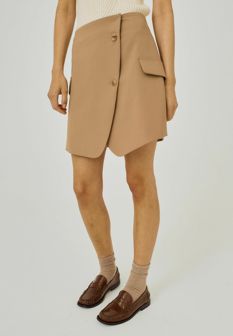 Beige omlottkjol med två knappar, sidofickor, slät textur och rundad nederkant, i kombination med bruna loafers och ljusbruna strumpor.