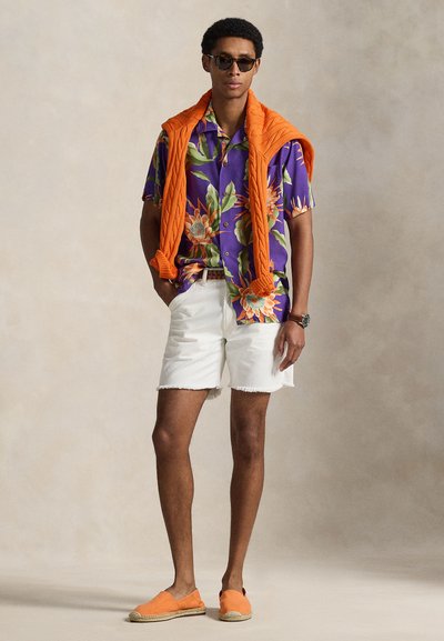 Mężczyzna w okularach przeciwsłonecznych, fioletowej koszuli w kwiaty, białych szortach, pomarańczowym swetrze narzuconym na ramiona, pomarańczowych wsuwanych butach, stoi z rękami w kieszeniach.