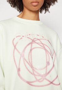 Gros plan d'une personne portant un sweat-shirt de couleur claire avec un motif abstrait en spirale rose et un texte pâle "Calvin Klein Jeans" sur la poitrine.