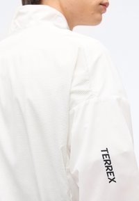 Personne en veste blanche avec un panneau arrière texturé et l'inscription « TERREX » en noir sur la partie supérieure de la manche, vue de dos à la hauteur des épaules.