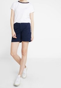 Bílý tričko s námořnickým a červeným pruhovaným límcem, spárované s námořnickými šortkami. Model má na sobě bílé tenisky. Jednoduchý, neformální a přiléhavý design.