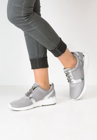 Szare buty sportowe z teksturowaną cholewką z materiału, metalicznymi akcentami i białymi gumowymi podeszwami. Z detalami takimi jak sznurowanie i boczne panele.