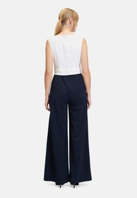 Combinaison sans manches avec un haut blanc et un pantalon large bleu marine. Comprend une fermeture éclair dans le dos et une taille ajustée, créant une silhouette élancée.