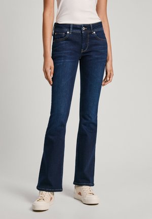 Femme portant un jean bootcut bleu foncé et des baskets beige, debout avec les bras détendus le long du corps devant un fond uni.