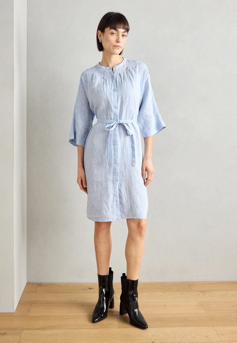 Svetlo modra lanena obleka s tremi četrtinami rokavi, sprednjimi gumbi in pasom za zavezovanje v pasu. Nosijo jo z črnimi škornji na peto do gležnjev.