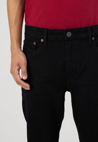 Los jeans de mezclilla negra presentan un corte ajustado, cierre de botón metálico y remaches de cobre en los bolsillos frontales. La tela tiene una textura con un aspecto robusto.