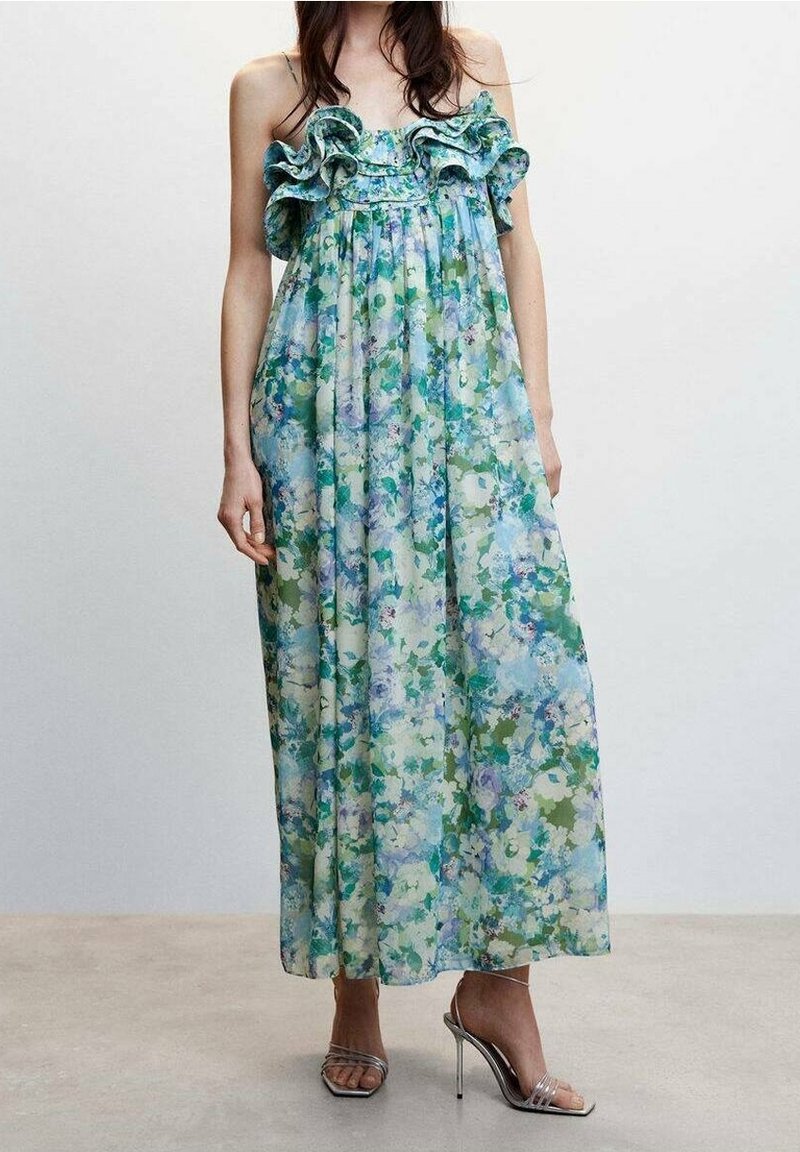 Femme portant une longue robe fleurie bleue et verte avec des volants au niveau de l'encolure et des sandales argentées à talons hauts.