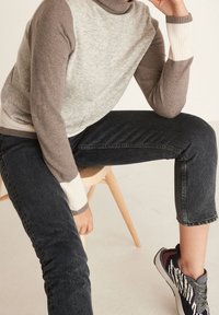 Grey and brown color-block turtleneck sweater with ribbed cuffs, paired with dark denim jeans; seated on a light wood chair.