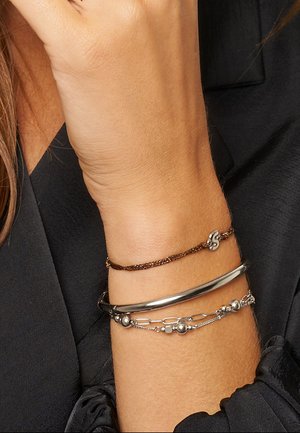 Drie armbanden om een pols: een dunne bruine ketting met een letteraccent, een gladde zilveren bangle en een zilveren armband met kralen.