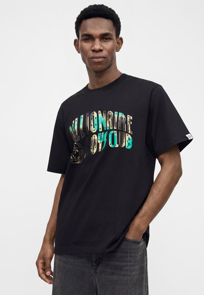 Νεαρός άνδρας φοράει μαύρο μπλουζάκι Billionaire Boys Club με πολύχρωμο κείμενο, με το ένα χέρι στην τσέπη, μπροστά σε ένα απλό φωτεινό φόντο.